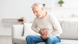 © aukkasit - Elderly Man Experiencing Severe Abdominal Pain Sitting on Sofa at Home