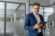 © Stock 4 You - Focused senior mature Indian or Latin entrepreneur businessman holding digital pc tablet standing in office. Smiling hispanic man in suit working using touchpad computer for business work project