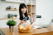 © Nuttapong punna - Asian woman working remotely on a laptop and tablet in a cozy kitchen workspace. Perfect for education, freelance