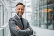 © Helena GARCIA - Hombre caucasico español, senior de mas de 50 años, mirando a cámara, sonriendo, llevando traje gris con corbata azul marina y camisa azul claro, sobre fondo luminoso de edificio de oficinas