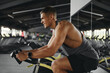 © Davidovici - Side view of handsome guy exercising on stationary cycle in modern gym while holding handle bar tightly and looking forward. Fitness and health care concept.