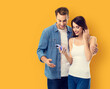 © vgstudio - Image - happy expressive surprise couple, family, man woman wear white t-shirt hold show cellphone smartphone mobile cell phone, isolate orange yellow background. Holiday sales credit money ad concept