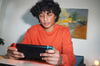 © Studio Marmellata - An Arab young man is focused while playing a handheld video game console. He is wearing an orange sweater and sitting at a table.