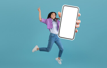 Excited asian lady showing empty smartphone screen while jumping up over blue studio background, collage, full body length, banner. Happy woman recommending cool mobile app or website