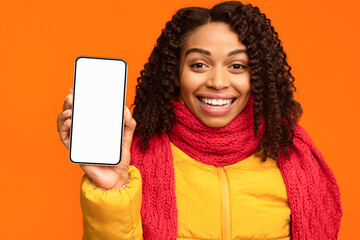 Poster - Joyful African Lady Showing Phone With Empty Cellphone Screen To Camera Approving Smartphone Standing Over Yellow Studio Background, Wearing Winter Jacket. Mobile Application Advertisement Concept