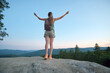 © bilanol - Sportive woman standing alone on mountainside trail with raised up arms. Female hiker enjoying view of evening nature from rocky cliff on wilderness path. Active lifestyle concept