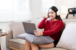 © NAMPIX - Video Conference. Smiling asian woman with headset having web call on laptop at home, talking at camera while sitting on sofa in living room