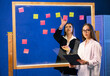 © Vadim - Team brainstorming session. Two women collaborate in a colorful brainstorming session, using sticky notes on a transparent board in a modern office.