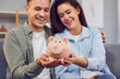 © Studio Romantic - Portrait of a happy young married couple planning their family budget sitting on sofa at home and holding piggy bank in hands saving money for future. Investments and financial freedom concept.