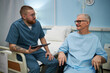 © DragonImages - Medical professional engaging senior patient wearing hospital gown while discussing health. Patient sitting on hospital bed, holding digital tablet in medical consultation setting