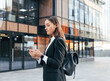 © Artem Varnitsin - Female in black formal clothes wearing backpack typing on mobile phone while standing against an office building