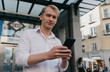 © BullRun - Confident young Caucasian businessman using smartphone outdoors, urban background, stylish casual outfit, symbolizing modern freelance work, digital finance, networking, success, and independence