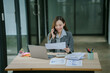 © PSG - Smiling asian businesswoman talking on phone and using laptop while working at office desk