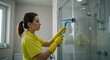 © SimpleDesignStudio - Young adult Caucasian woman in a bright yellow shirt and protective gloves meticulously cleaning a glass shower door in a modern bathroom using a squeegee tool.