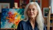 © ULTRA STOCK'S - Female painter from the baby boomer generation with long, white hair posing in front of her paintings