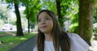 © Marco - Young girl with long hair gazing upwards in a tree-lined park, wearing a white blouse, looking thoughtful and hopeful, surrounded by lush greenery on a sunny day