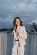 © SHOTPRIME STUDIO - Confident woman in light business attire poses on a bridge against a skyline backdrop, exuding professionalism and modernity in an urban setting.