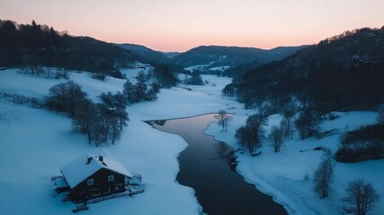 Poster - Aerial view of a serene winter landscape featuring a snow-covered valley with a winding river and a small cabin. The sky displays soft hues of pink and orange during the sunset