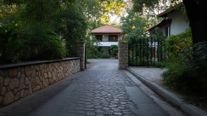 Poster - Pathway leading to a residential property with stone walls gate and houses with surrounding greenery. The scene showcases a serene suburban setting during daylight