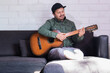 © Rodrigo - man in cap smiles while practicing guitar at home
