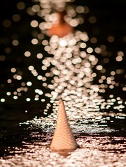 Poster - Close up view of a textured, light colored cone shaped object sitting on a dark, reflective surface. The background is blurred, showing a stream of bright bokeh lights suggesting water droplets or