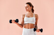 © HBS - A young woman with beautiful abs and a toned body, wearing white fitness attire, is lifting black dumbbells while standing in front of a soft peach-colored background in a studio.