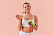 © HBS - A smiling, fit young woman in white fitness clothing eating a plate of salad against a soft peach-colored background in a studio.