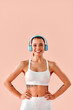 © HBS - A vertical image of a young woman in fitness attire with blue headphones, looking at the camera and smiling against a light pink background. A sporty lifestyle.