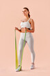 © HBS - Vertical image – A fit young woman in white sportswear working out with a green resistance band against a peach background. Fitness, motivation, healthy lifestyle.