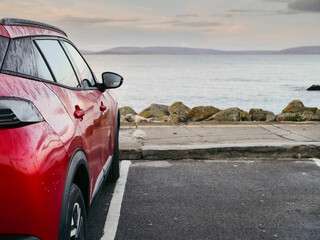 Wall Mural - Red car parked in car park with stunning view on nature scenery with ocean and mountains and cloudy sky. Silverstrand area, Galway city, Ireland. Rent motor for a tourist trip concept.