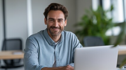 Fototapete - A happy business man sits at a well-organized desk in a contemporary office. He is working on his laptop and smiling, showcasing a positive work atmosphere in a bright environment
