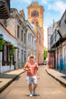 © Itza - Young Latino man in coral shirt, white shorts, and straw hat confidently walks through a colonial street in Cartagena, Colombia, enjoying a refreshing drink under the sun.