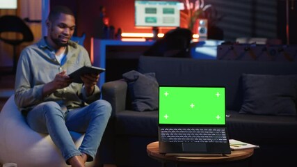 Poster - young male person enjoys leisure time next to chroma key on bean bag, uses his smartphone to watch a