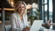 © Adi - happy middle-aged businesswoman using a laptop in an office setting, smiling while working
