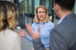 © Dexon Dee - Upset young woman in business attire raising hands in frustration while having a heated discussion with colleagues in a modern business district. Workplace disagreement and professional stress concept