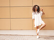 © halayalex - Beautiful smiling model with afro curls hairstyle dressed in summer hipster white dress.Sexy carefree female posing in street near yellow wall in sunglasses.Positive woman having fun