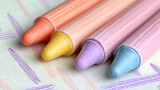 Close-up of four pastel colored crayons resting on a patterned surface.  Crayons are orange-peach, yellow, light purple, and light blue.  The crayons have a conical shape and are smooth.  
