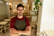 © we.bond.creations - Confident restaurant owner smiling with arms crossed, wearing an apron, posing in his modern restaurant dining room