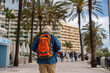 © sutulastock - An elderly man with a white beard and a cap, wearing a denim shirt and carrying a bright orange backpack, strolls along a palm tree-lined promenade on a sunny day
