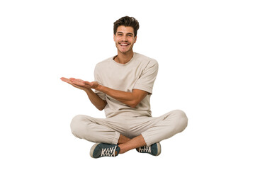 young caucasian man sitting on the floor isolated on white background holding a copy space on a palm