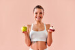 © HBS - A smiling, sporty young girl in a white top holds a green apple in one hand and a beautiful, high-calorie dessert in the other. A choice and balance in nutrition.