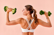 © HBS - A young woman in a white sports bra, seen from the back, holds fresh broccoli like dumbbells while doing a triceps exercise. A creative advertisement for healthy food and fitness.