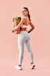 © HBS - A vertical image of a fitness trainer in white sportswear holding a bag of vegetables and healthy food, standing against a soft peach background. Healthy eating and fitness.