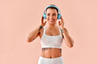 © HBS - A happy young woman in a white sports bra and leggings is smiling while holding her hands near her blue headphones, against a soft peach background in a studio. Living a sporty lifestyle.