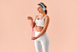 © HBS - Woman in white fitness wear and blue headphones doing an arm stretch exercise with a resistance band against a soft peach background.Training motivation using supportive