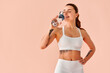 © HBS - A beautiful young woman in white fitness wear drinking water from a bottle against a soft peach studio background.Copy space.