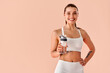 © HBS - A confident, beautiful adult woman wearing a white sports bra and leggings, holding a fitness water bottle and smiling against a soft peach studio background. Copy space.