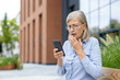 © Liubomir - An older woman is looking at her phone with a look of concern on her face, outdoors. She is holding her hand to her mouth as well.