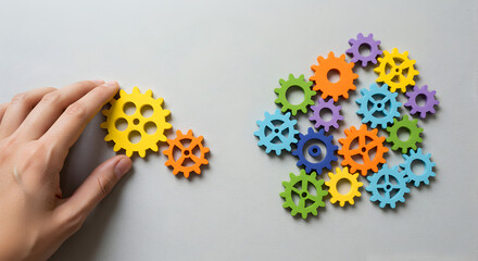 Poster - Hand Connecting Yellow and Orange Gear Pieces with Multiple Colorful Gears on Grey Background Representing Engineering or Teamwork