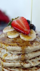 Wall Mural - Pouring maple syrup on top of oatmeal pancakes with fresh fruit. Vertical video. Close-up. Sweet dessert of breakfast food preparation.